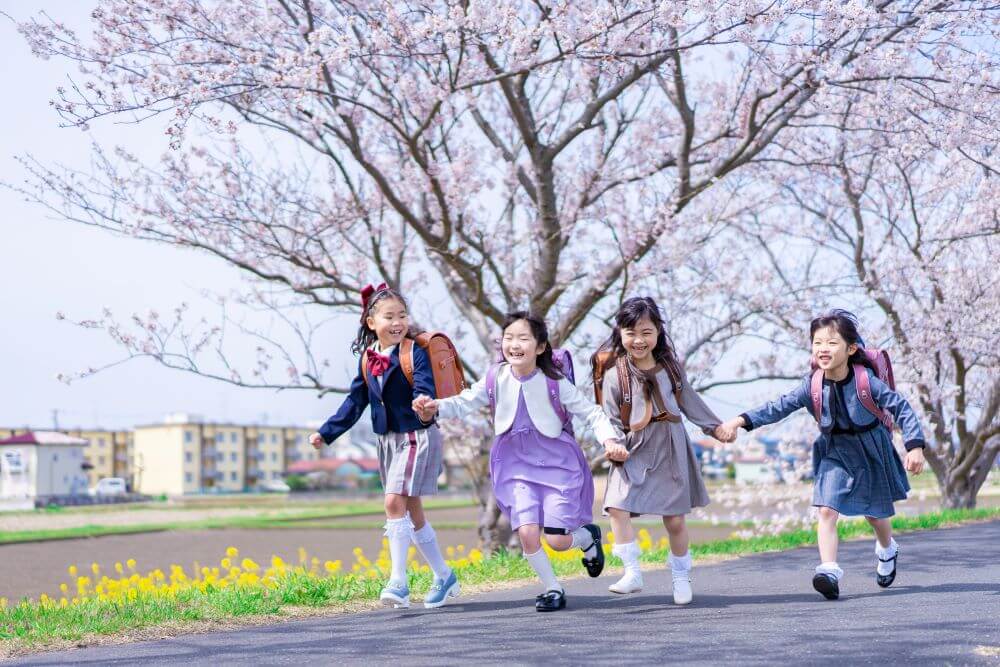 桜が並ぶ道で小学生が仲良く手を繋いで走っている